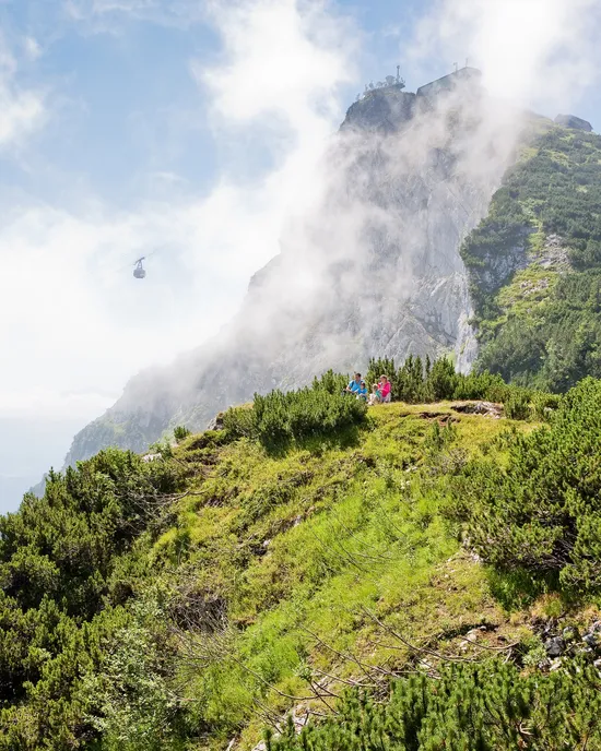 Escursionisti in pausa su sentiero alpino verde con funivia e cima montuosa avvolta dalla nebbia